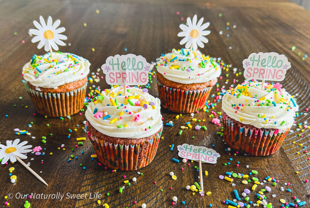 Four beautifully decorated carrot cake cupcakes with sugar-free frosting and spring-themed toppings, displayed on a light, airy surface with seasonal touches like pastel colors and fresh florals.