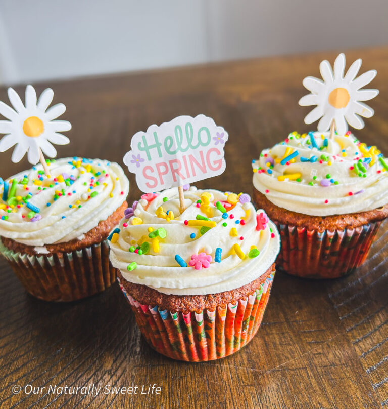 Three spring-themed carrot cake cupcakes decorated with sugar-free cream cheese frosting and pastel sprinkles, arranged on a light, rustic surface with soft floral accents.