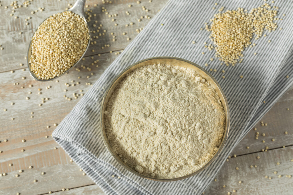Top view of a bowl filled with light-colored quinoa flour placed on a striped cloth, with raw quinoa seeds scattered around and a spoonful of whole quinoa nearby on a rustic wooden surface.